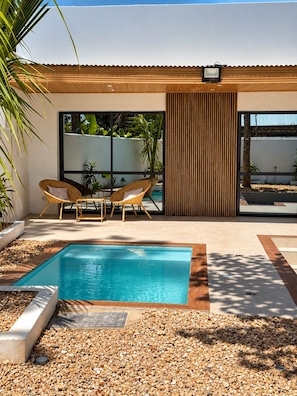 A modern outdoor pool area with a rectangular pool, surrounded by pebbles and a wooden deck. There are two wicker chairs and a table with cushions.