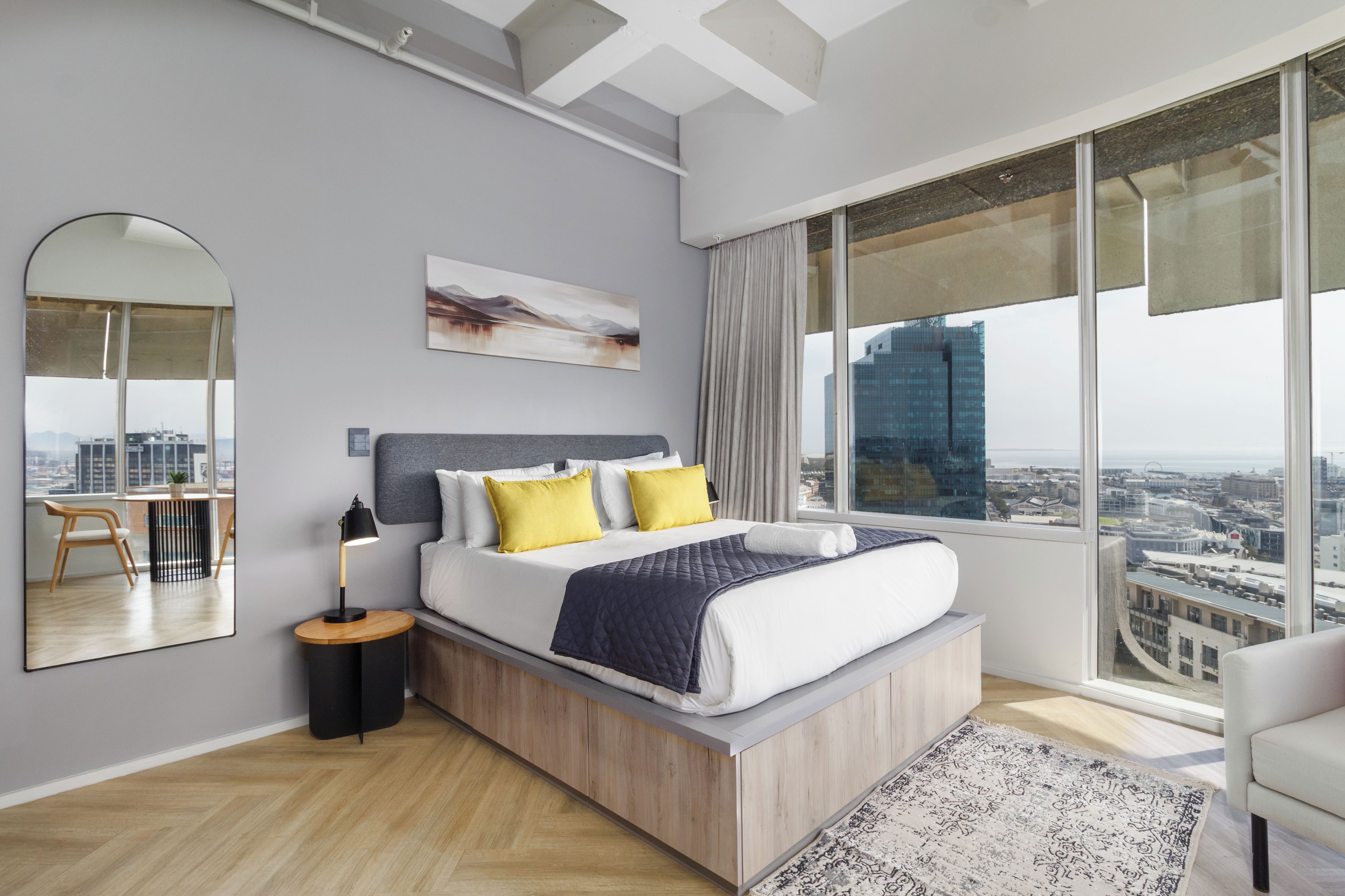 Une chambre moderne avec un grand lit, une table de chevet, une lampe, un miroir et une vue sur la ville.
