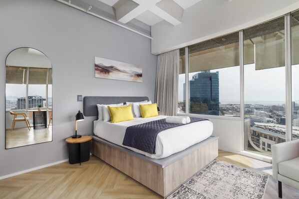 A modern bedroom with a large bed, a bedside table, a lamp, a mirror, and a city view. - Central CBD Apartment | Beautiful Views (Cape Town)