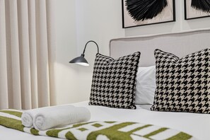Ein ordentlich bezogenes Bett mit einem aufgerollten weißen Handtuch, zwei Kissen mit Hounds-Tooth-Muster, einer schwarzen Schreibtischlampe und einer Decke in Grün-Weiß.