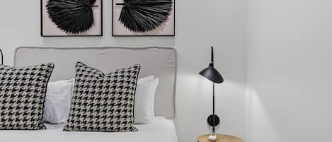 Un dormitorio con una cama, dos obras de arte en la pared, una lámpara de noche y una pequeña mesa de madera.