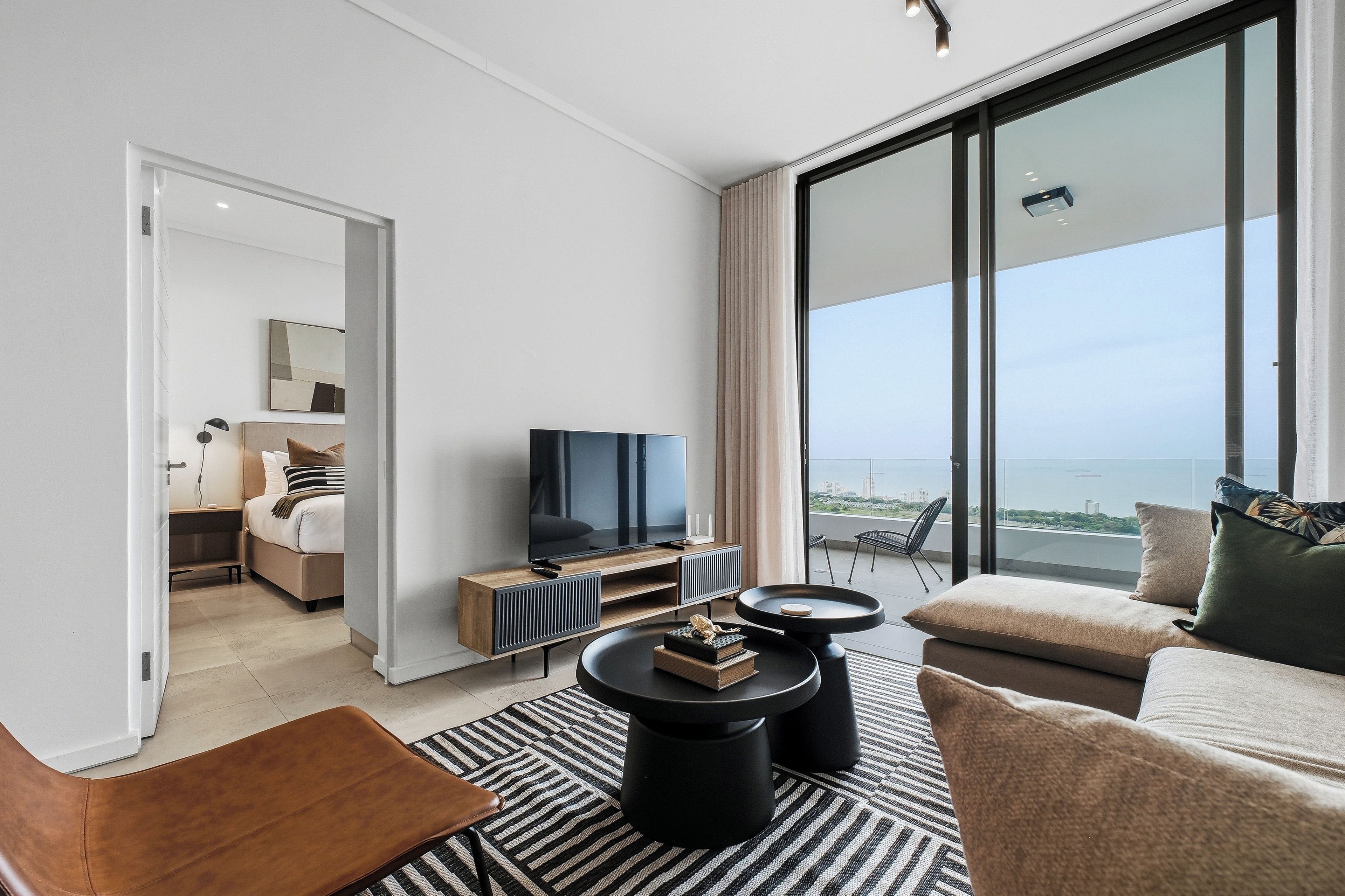 A modern hotel room with a balcony, a flat-screen TV, a sofa, and a dining area.
