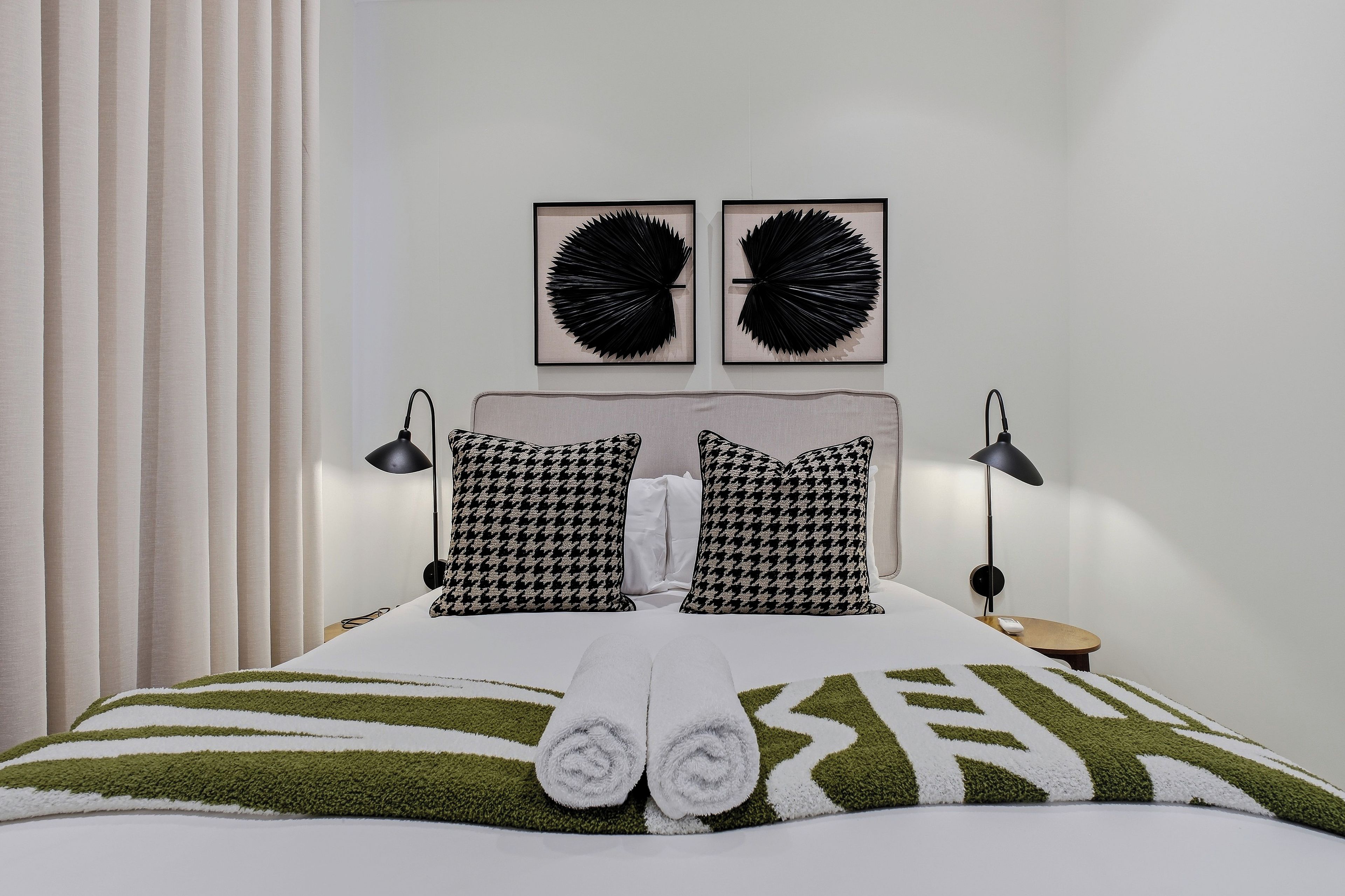 Ein ordentlich bezogenes Bett mit einer grün-weißen Streifendecke, zwei Hundepunkt-Kissen und je eine schwarze Lampe auf beiden Seiten.
