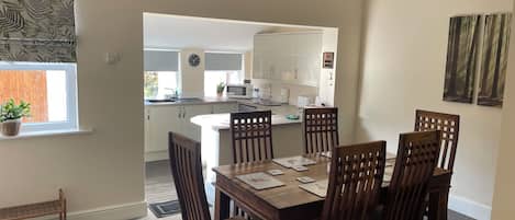 A dining area with a wooden table and chairs, a hanging light fixture, and a view into the kitchen.