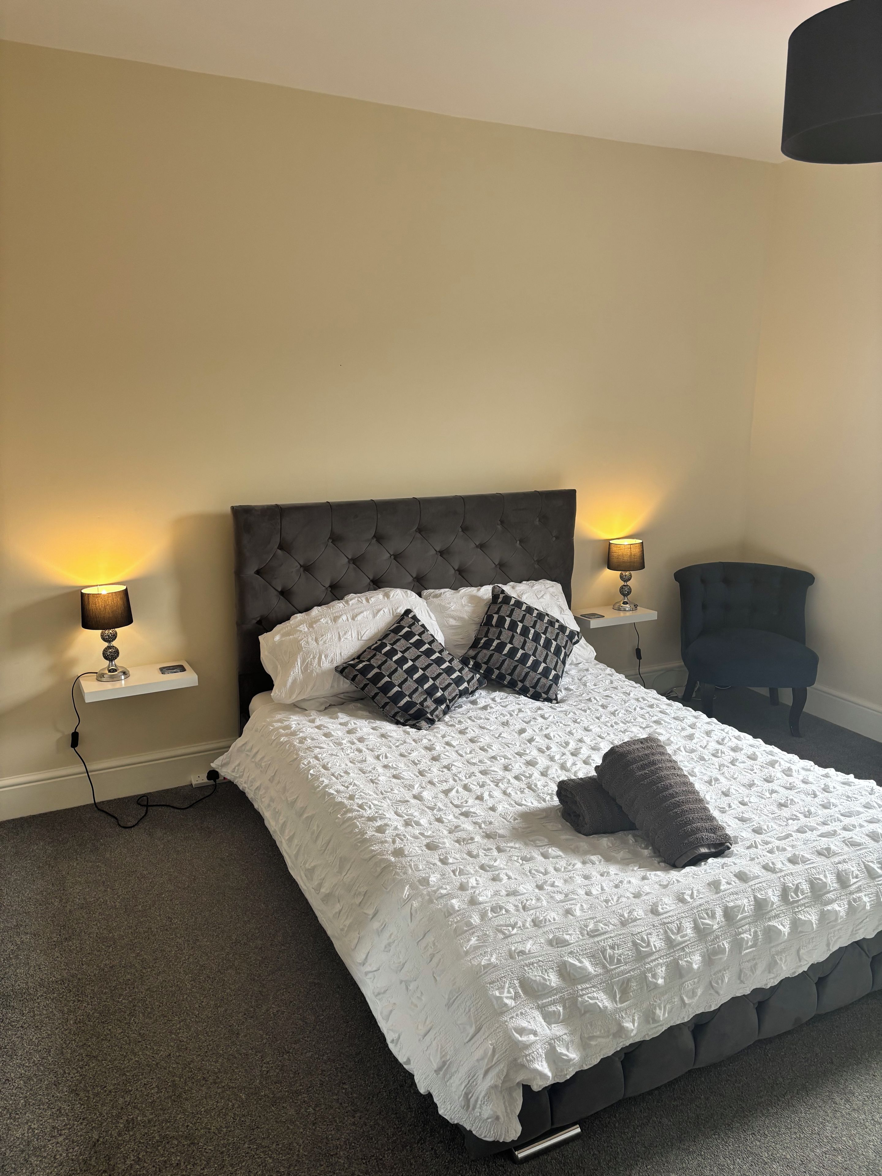 A neatly made bed with a white textured bedspread and two patterned pillows, flanked by two bedside lamps on white nightstands.