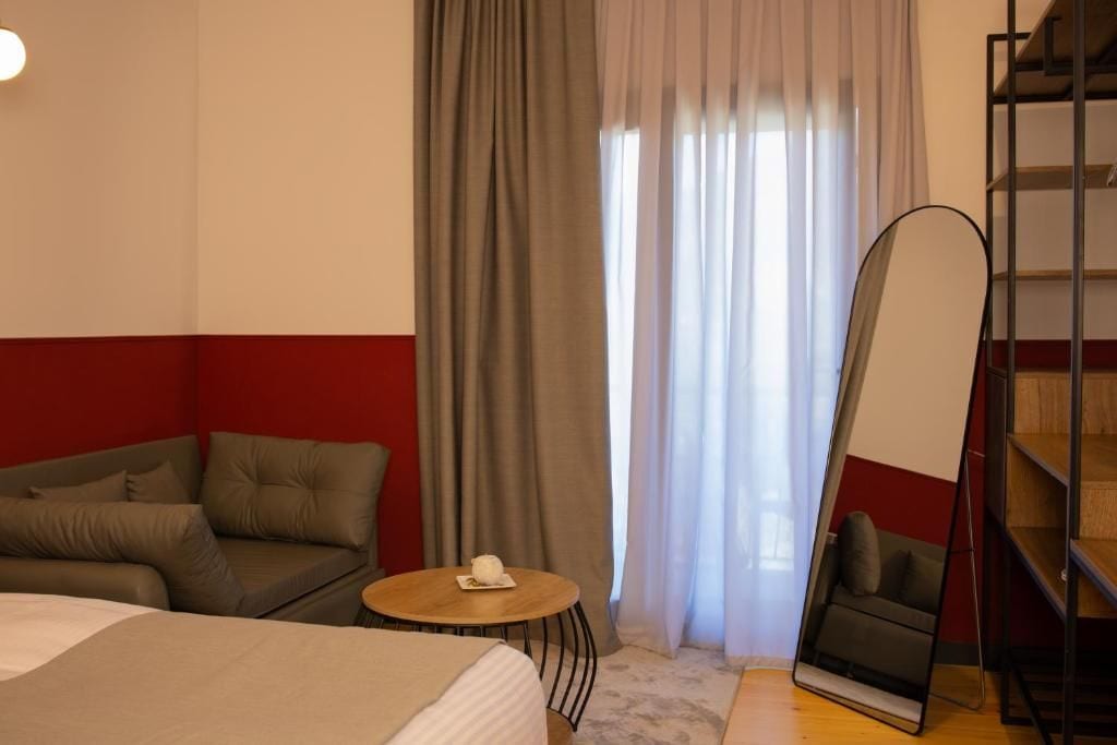 Habitación de hotel con cama, sofá, una pequeña mesa redonda con un control remoto, un espejo grande y una estantería.