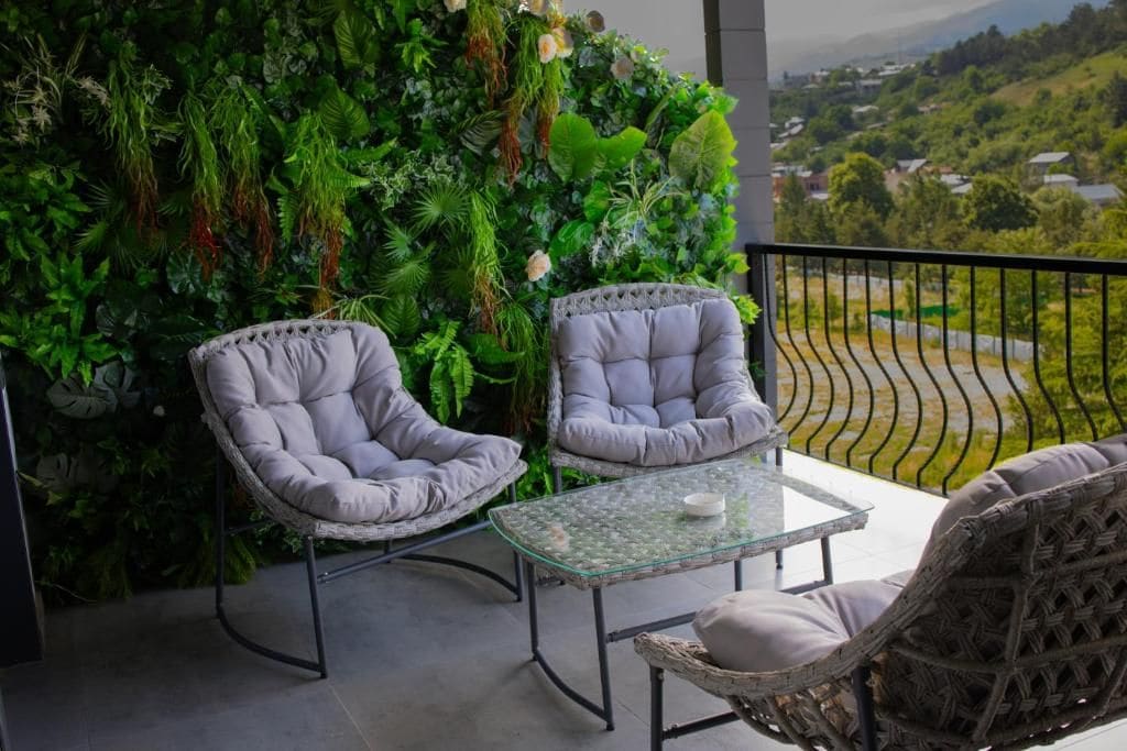 Un balcón con dos sillas acolchadas, una mesa de vidrio y una pared verde exuberante.