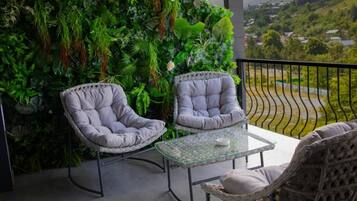 Un balcón con dos sillas acolchadas, una mesa de vidrio y una pared verde exuberante.