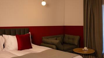 A hotel room with a bed, a red and white headboard, a green armchair, a round wooden side table, and a single hanging light fixture.