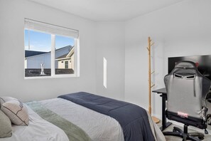 A bedroom with a bed, a desk, a chair, a window with a view of a house, and a coat rack. - Golden Sage Gathering House | Pet-Friendly 4BR Nampa Home • Sleeps 10 • Gym • Fenced Yard (Nampa)