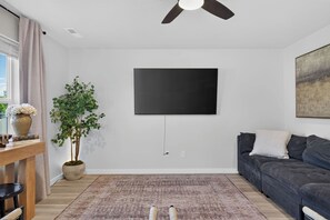 A modern living room with a flat-screen TV mounted on the wall, a dark-colored sofa, a wooden coffee table with books and a vase of flowers, a potted plant, and a patterned rug. - Golden Sage Gathering House | Pet-Friendly 4BR Nampa Home • Sleeps 10 • Gym • Fenced Yard (Nampa)