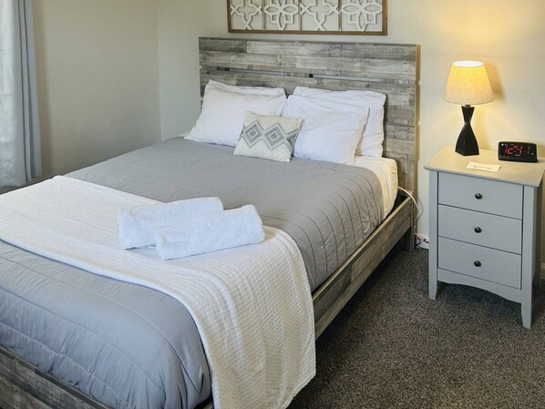 A neatly made bed with a grey and white duvet, a matching pillow, and a folded towel on the bed. There is a bedside table with a lamp and a digital clock. - Morning Star Cottage (Star)