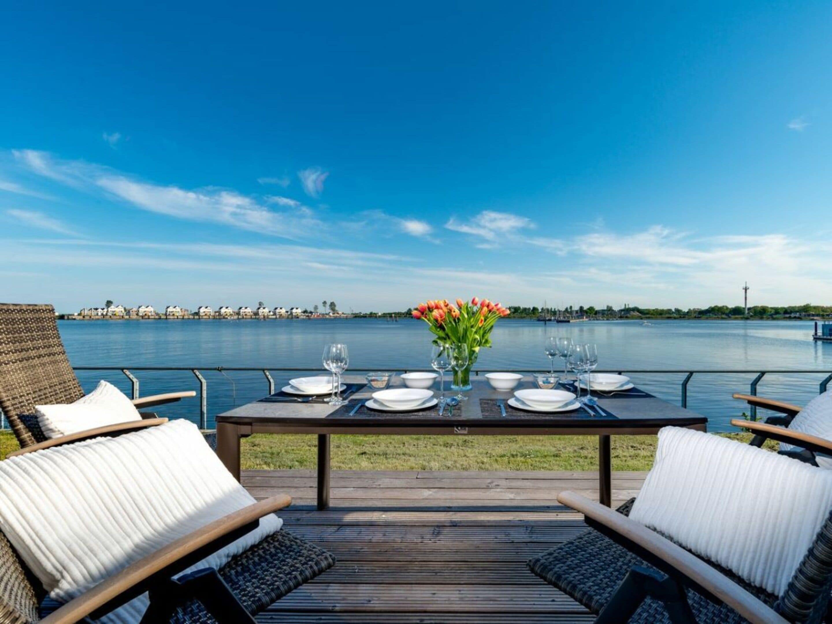 An outdoor dining area with a view of a body of water, a table set for two, and a row of chairs.