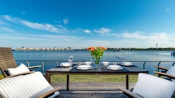 An outdoor dining area with a view of a body of water, a table set for two, and a row of chairs.