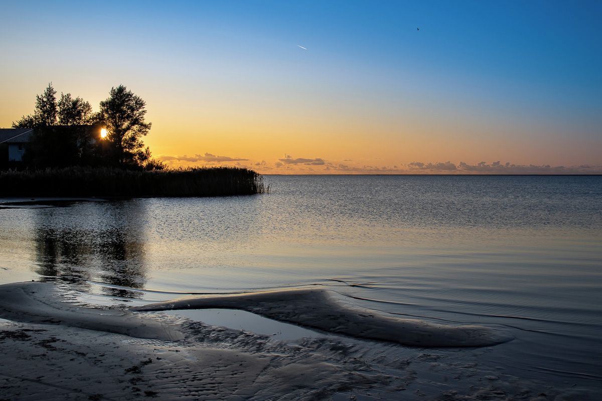 A calm body of water reflecting the sky during sunset, with a house and trees on the left.