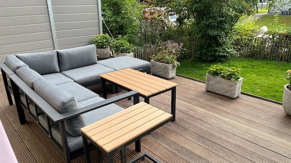 A modern outdoor seating area with a grey sectional sofa, two wooden tables, and potted plants.