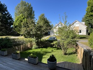 A residential area with a wooden deck, potted plants, a car parked on the street, and houses in the background.