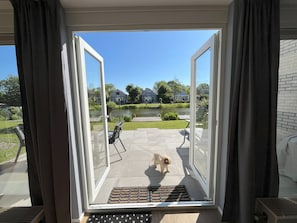 A view from a room with an open sliding glass door to a patio with a small dog, a pool, and houses in the background. - Modern vacation home with a garden (fenced on the west side) and a private dock on the IJsselmeer (Makkum)