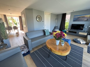 A modern living room with a grey sofa, a wooden coffee table with a vase of flowers, a flat-screen TV, and a large window with curtains.