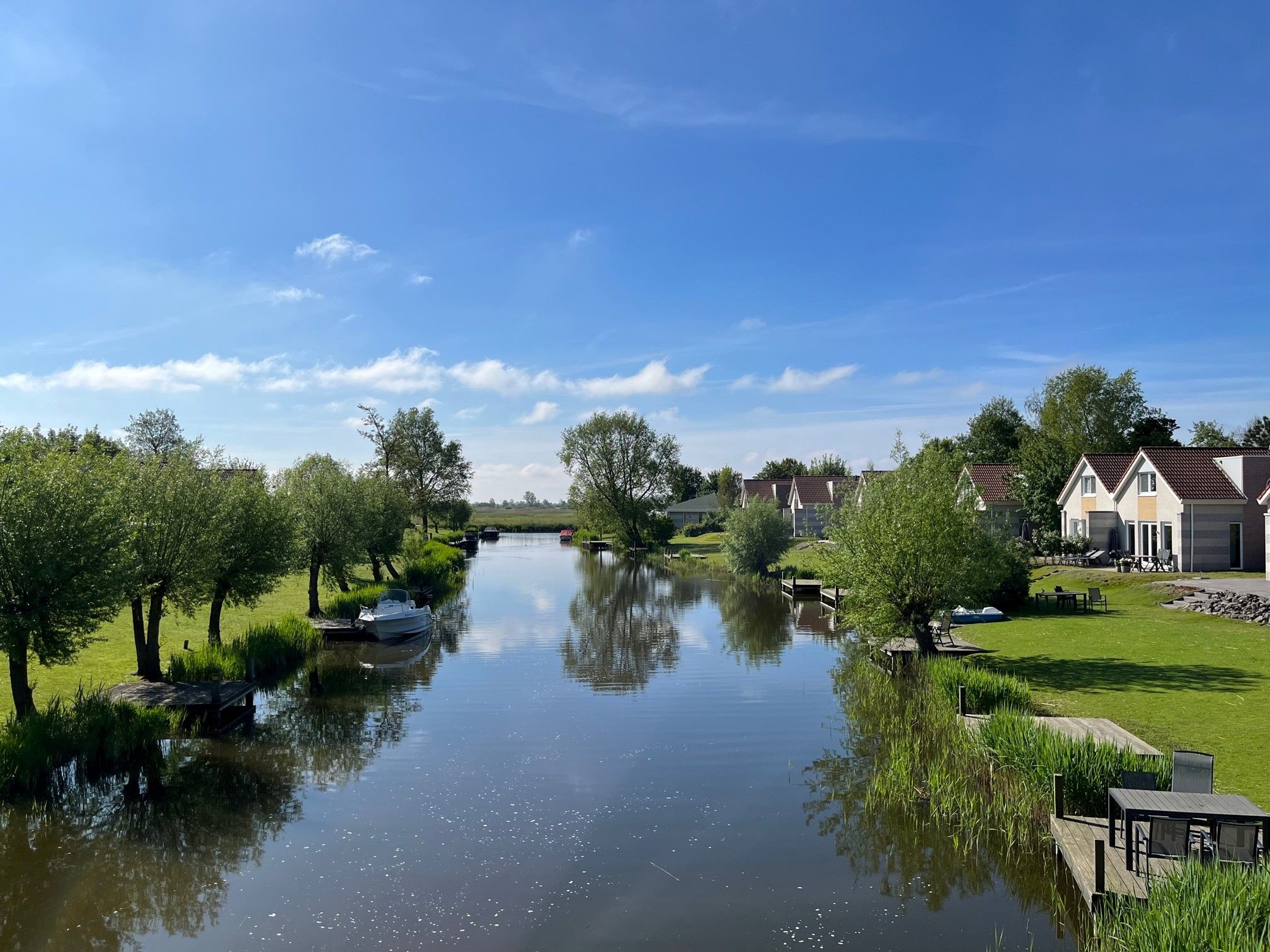 Een rustige rivier met een houten steiger, omgeven door groen en moderne huizen onder een heldere lucht.