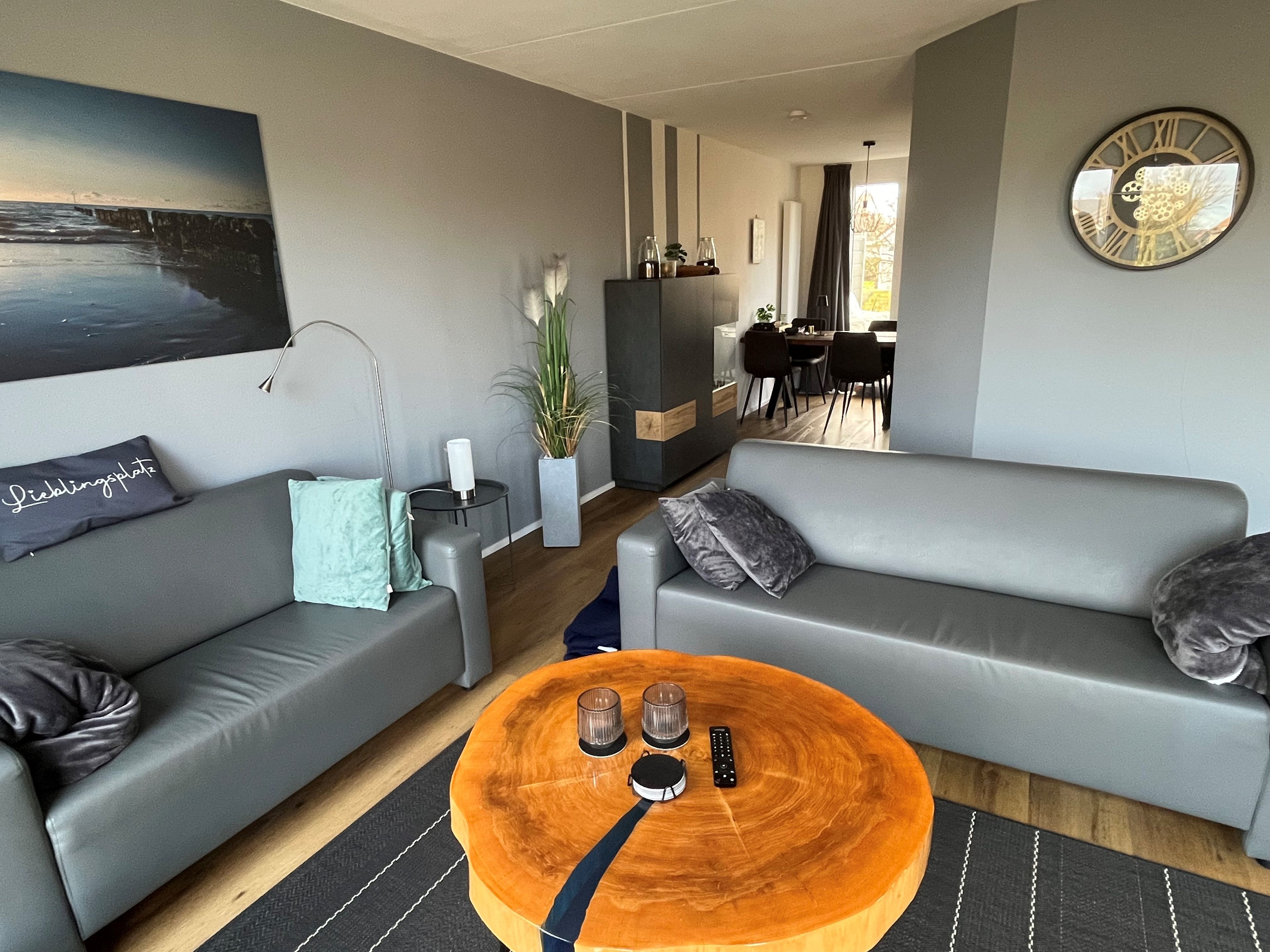 A modern living room with two grey sofas, a wooden coffee table with remote controls, and a dining area in the background.