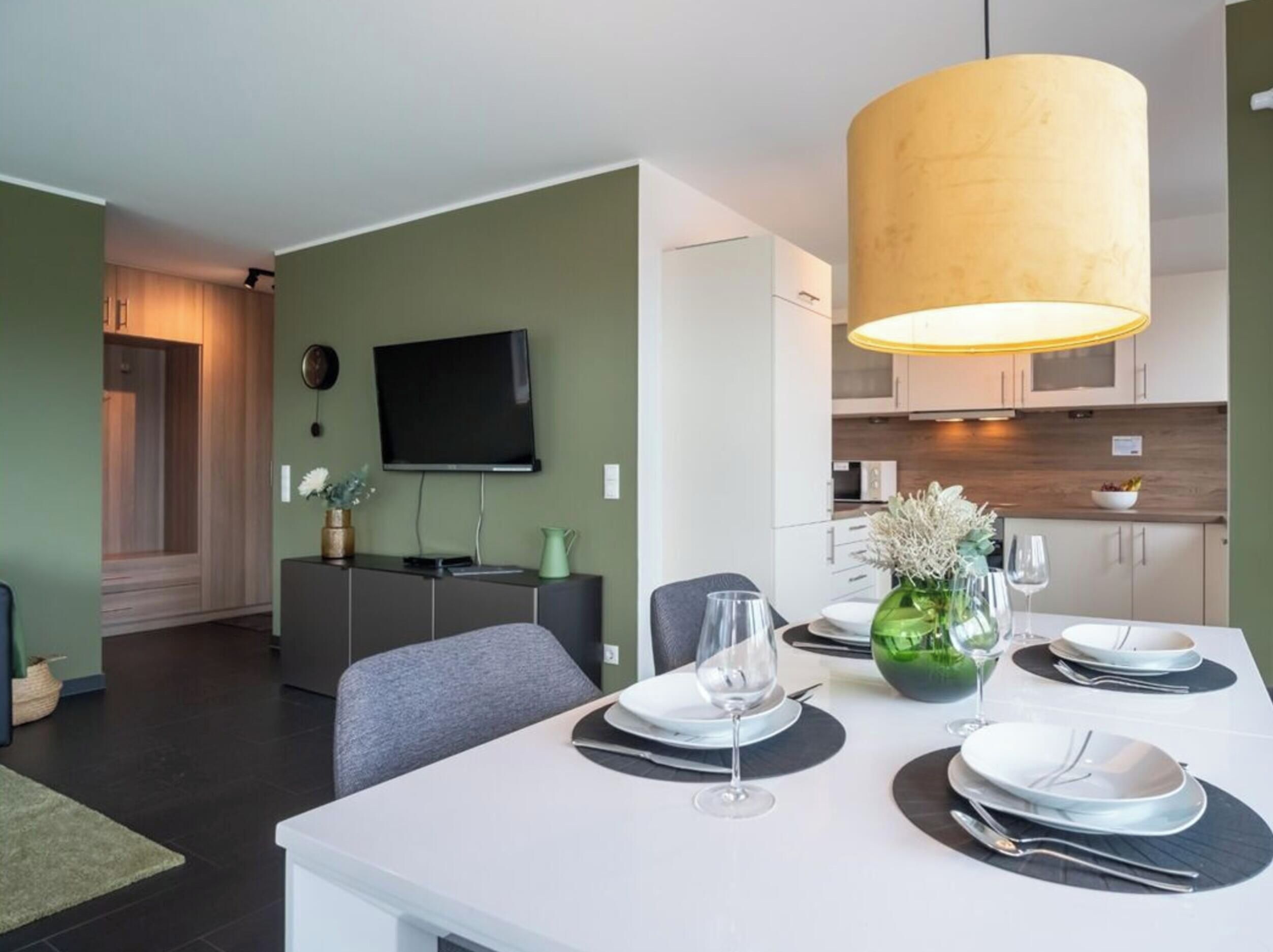 A modern dining area with a table set for two, a television mounted on the wall, and a kitchen in the background.