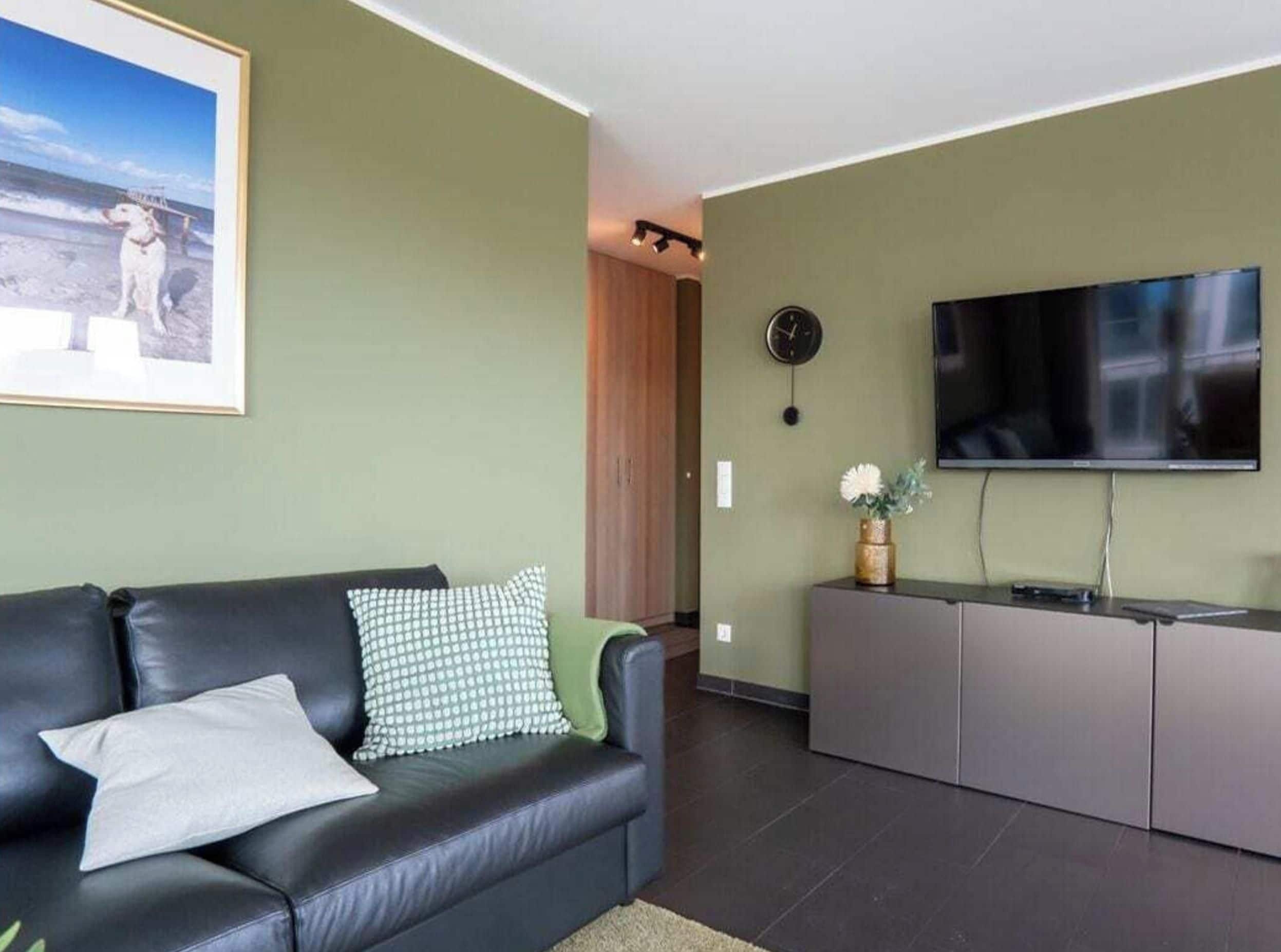Un salon avec un canapé en cuir noir, un téléviseur à écran plat fixé au mur et un tableau encadré au-dessus du canapé.