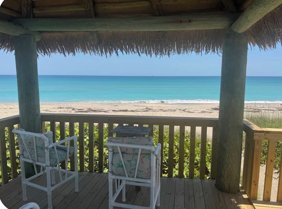 A wooden deck with two white chairs and a table, overlooking a beach with clear blue water.