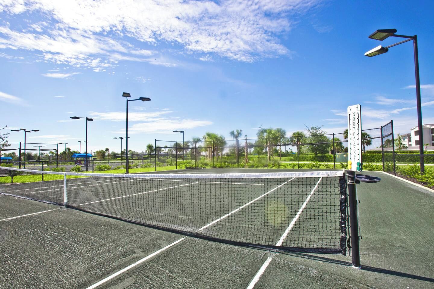 Un court de tennis avec un filet, entouré d’une clôture, et des lampadaires.