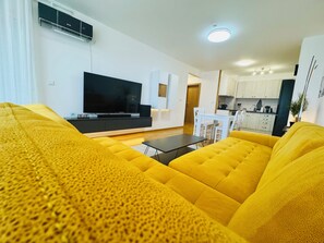 A modern living room with a large sofa, a flat-screen TV, and a dining area with a white table and chairs.