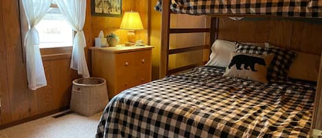 Ein Zimmer mit einem Etagenbett, einem Nachttisch mit Lampe, einer kleinen Kommode und einem Fenster mit transparenten Vorhängen.