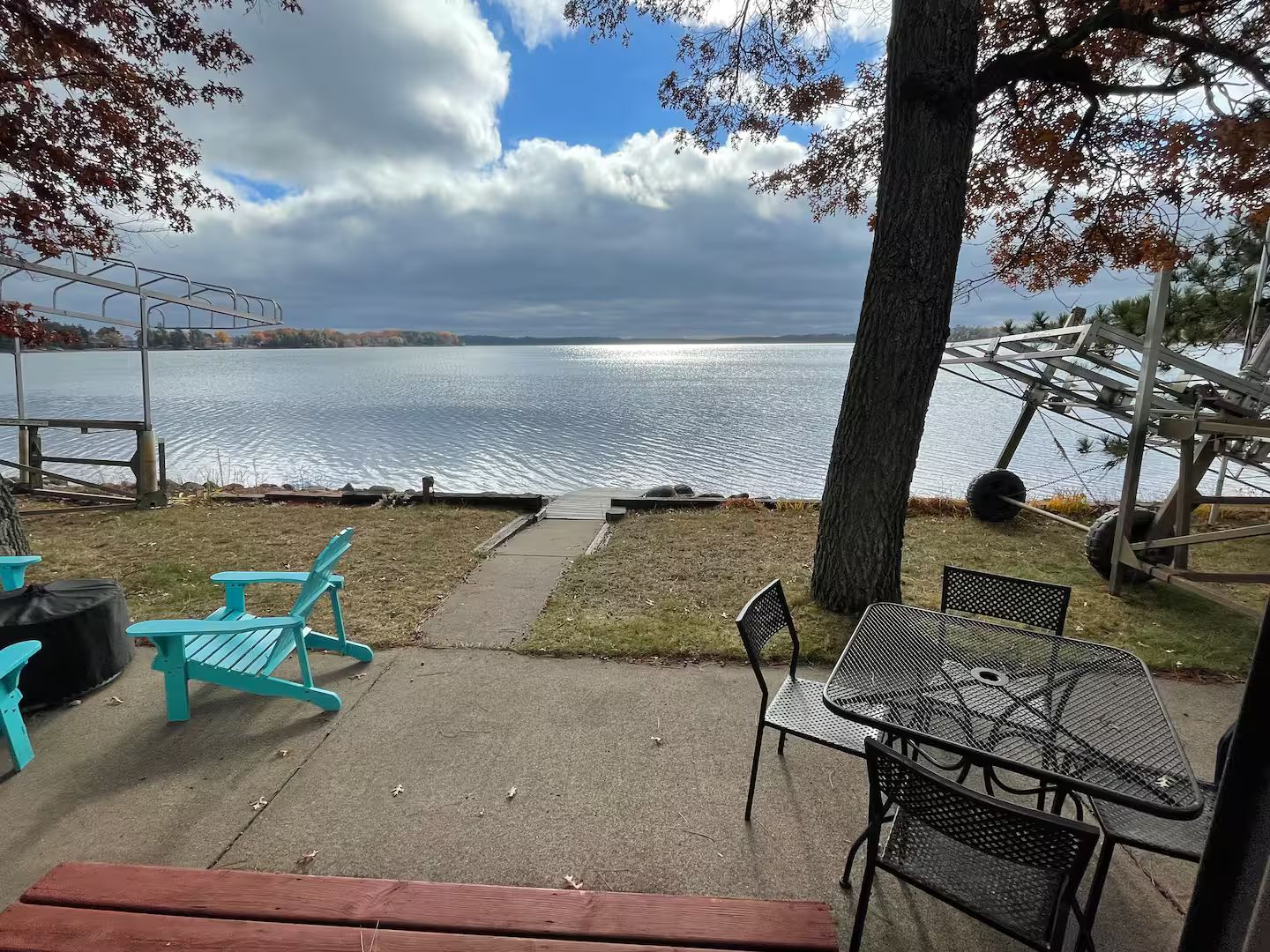 Un espacio de descanso junto al lago con una mesa y sillas de metal, un banco de madera y vistas al lago.