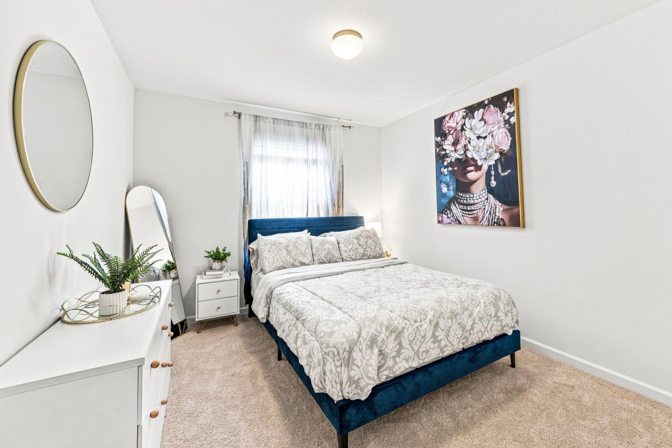 A bedroom with a bed, a bedside table, a mirror, a window with curtains, and a wall-mounted picture.