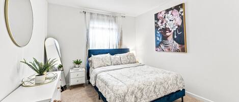 A bedroom with a bed, a bedside table, a mirror, a window with curtains, and a wall-mounted picture.