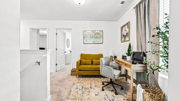 A hallway leading to a living room with a mustard sofa, a grey armchair, a wooden desk, and a map artwork on the wall.