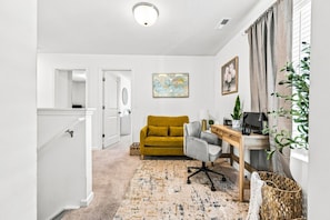 A hallway leading to a living room with a mustard sofa, a grey armchair, a wooden desk, and a map artwork on the wall.