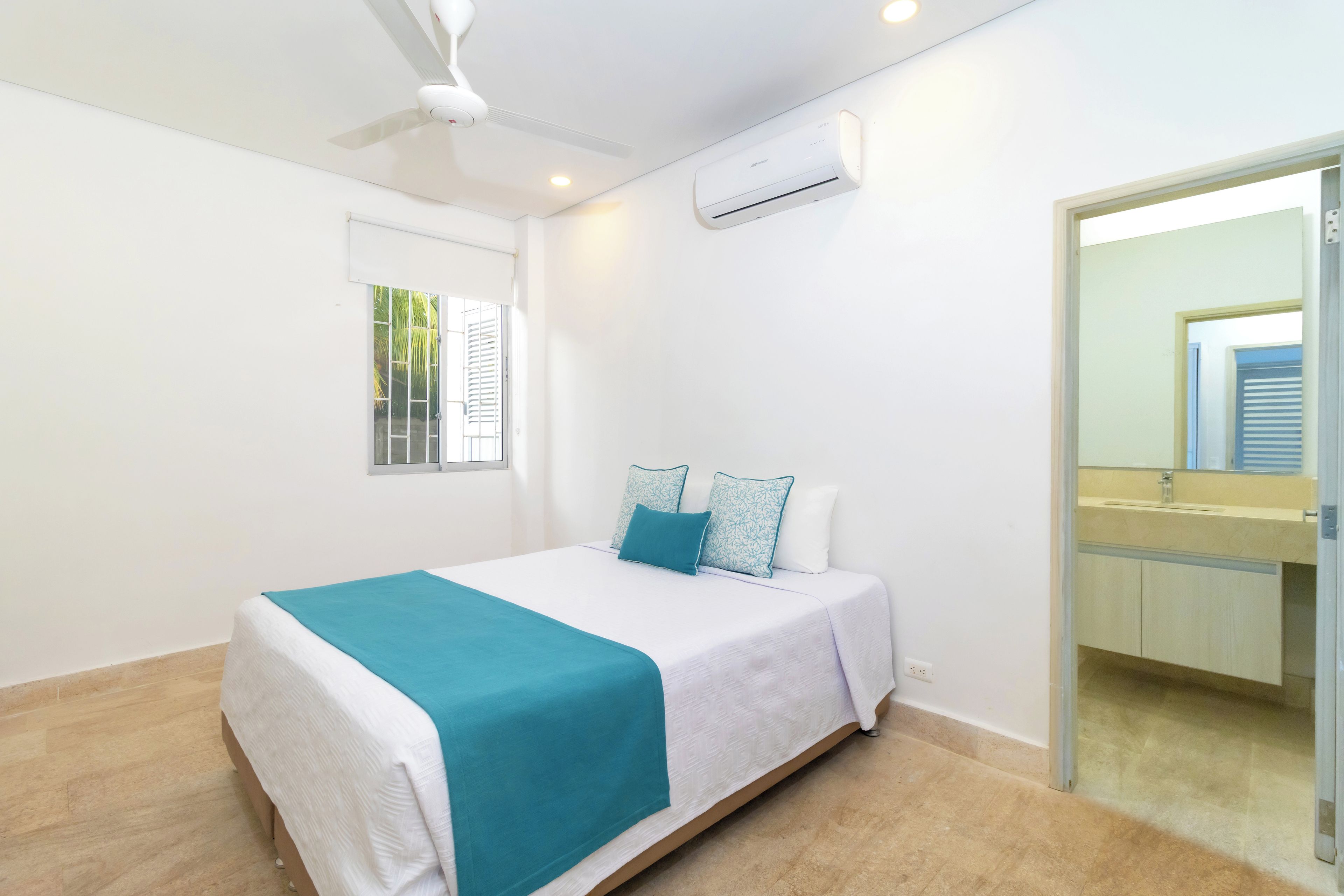 Un dormitorio con cama, ventilador de techo, unidad de aire acondicionado, ventana con persianas y un baño visible a través de una puerta abierta.