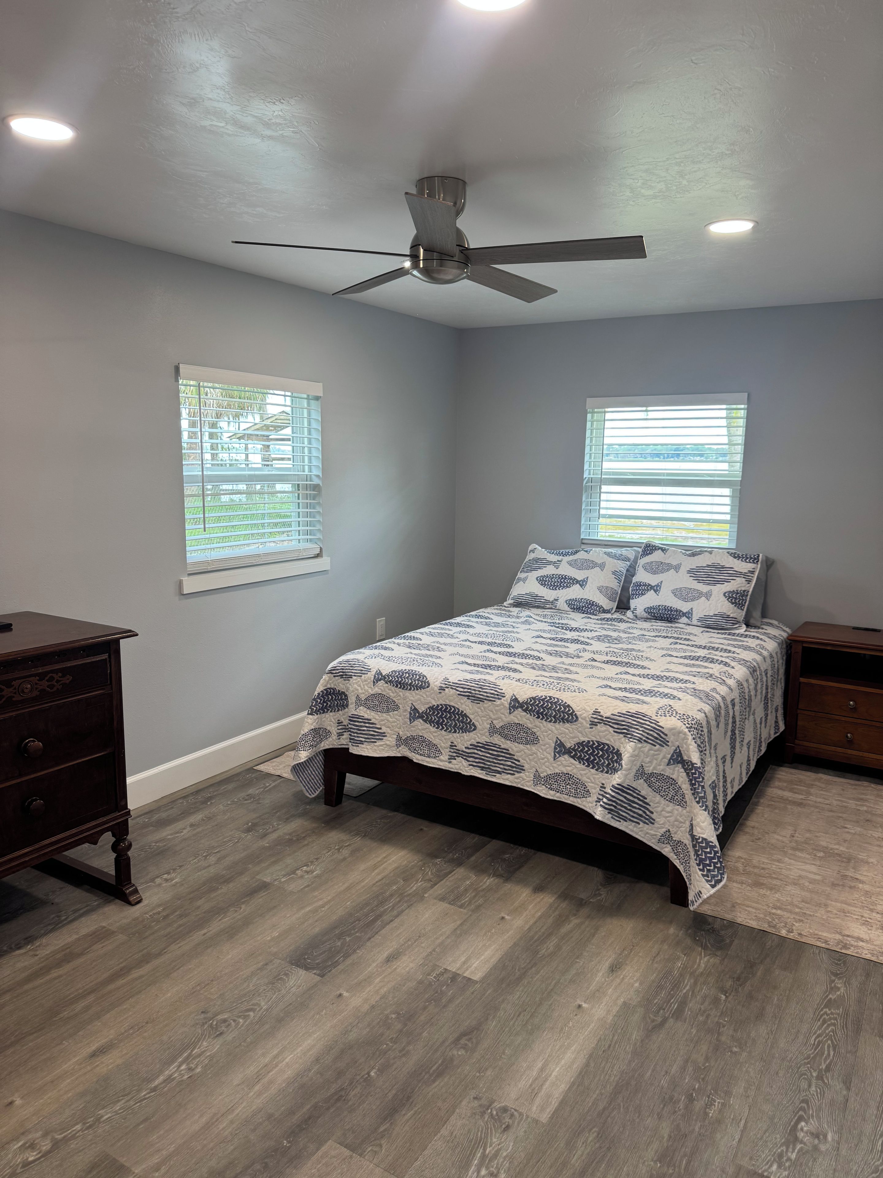 Quarto com cama, duas mesas de cabeceira, cômoda, ventilador de teto e duas janelas com persianas.