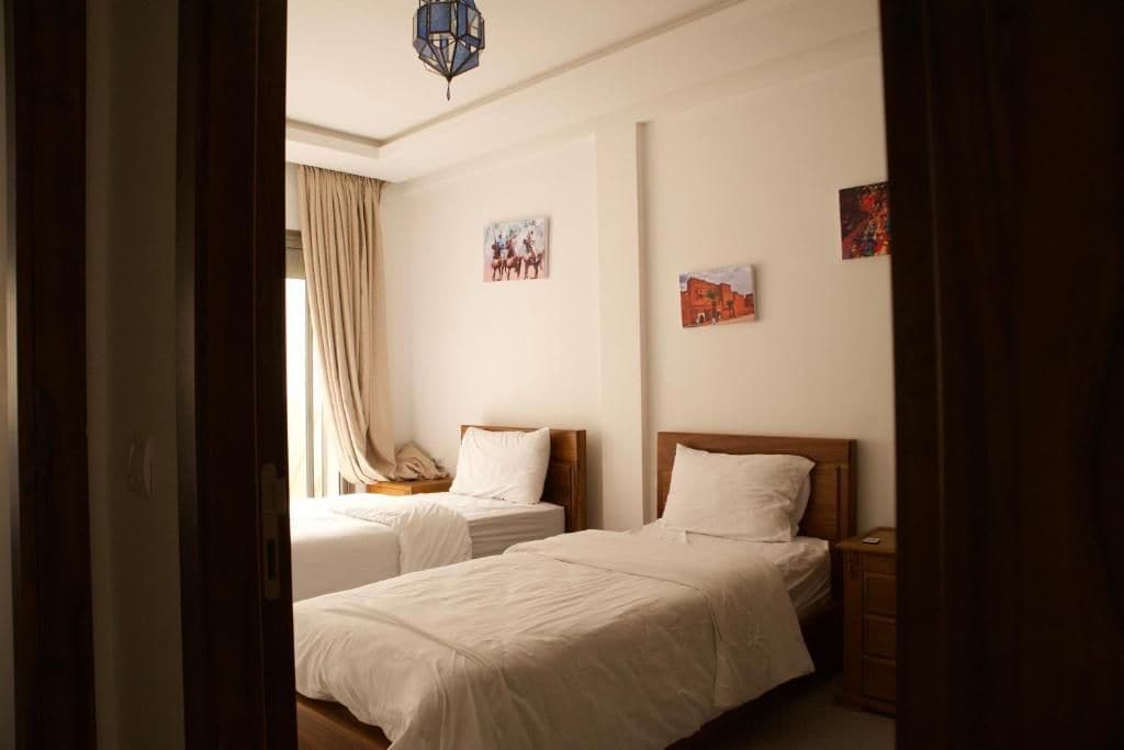 A hotel room with two beds, a wooden headboard, a nightstand, and a wall-mounted lamp.