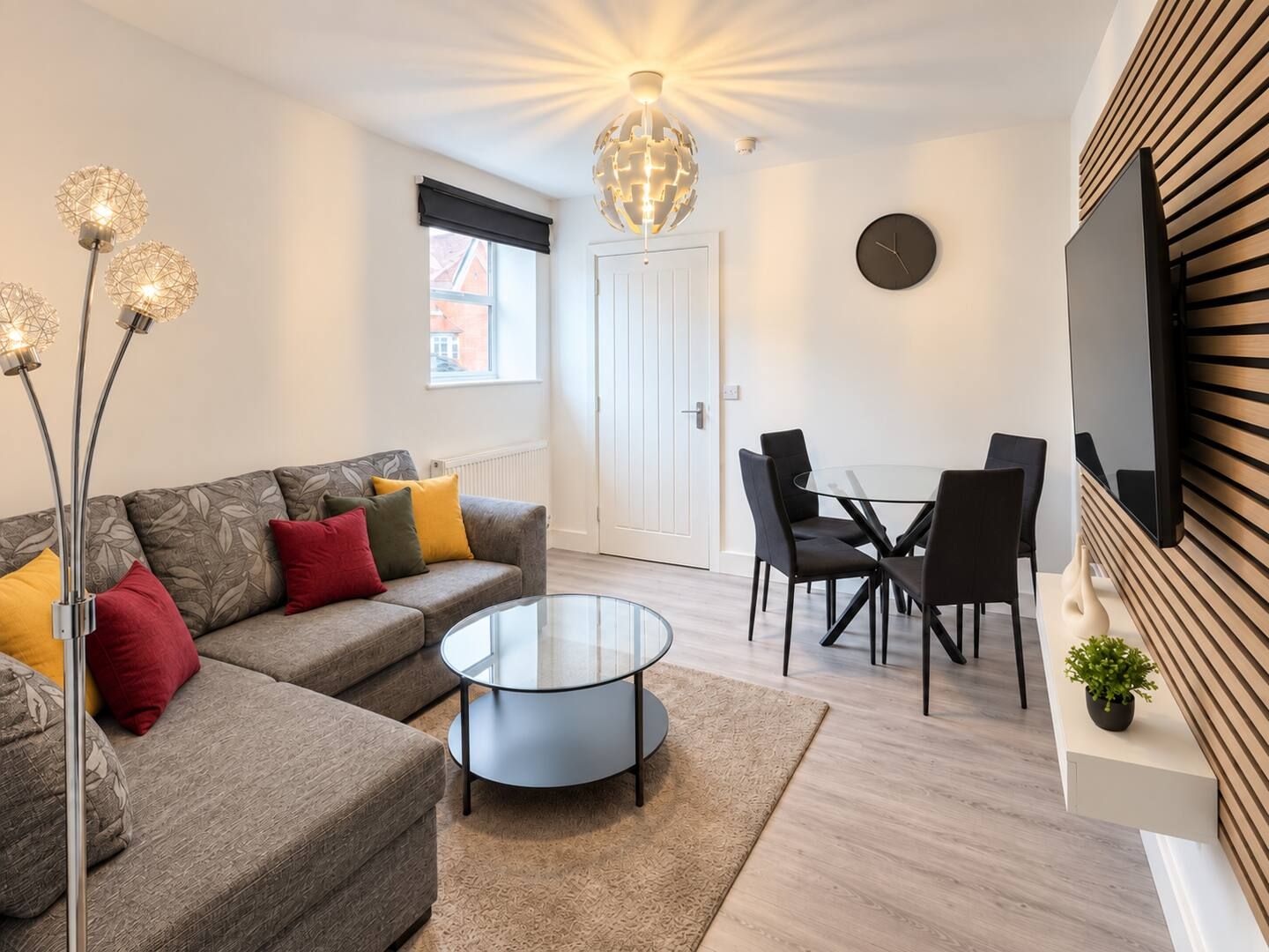 Een moderne woonkamer met een bank, salontafel, eettafel met stoelen, een aan de muur bevestigde televisie en een decoratieve gloeilamp in de vorm van een wereldbol.