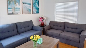 Un salón con dos sofás grises, una mesa de centro de madera con flores, y tres obras de arte con temática playera.