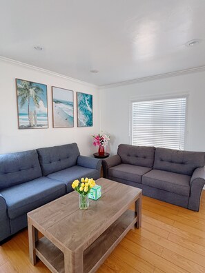 A living room with two grey sofas, a wooden coffee table with flowers, and three beach-themed wall art pieces. - 2BR Home Near Disneyland & LA - Free Parking (pico Rivera)