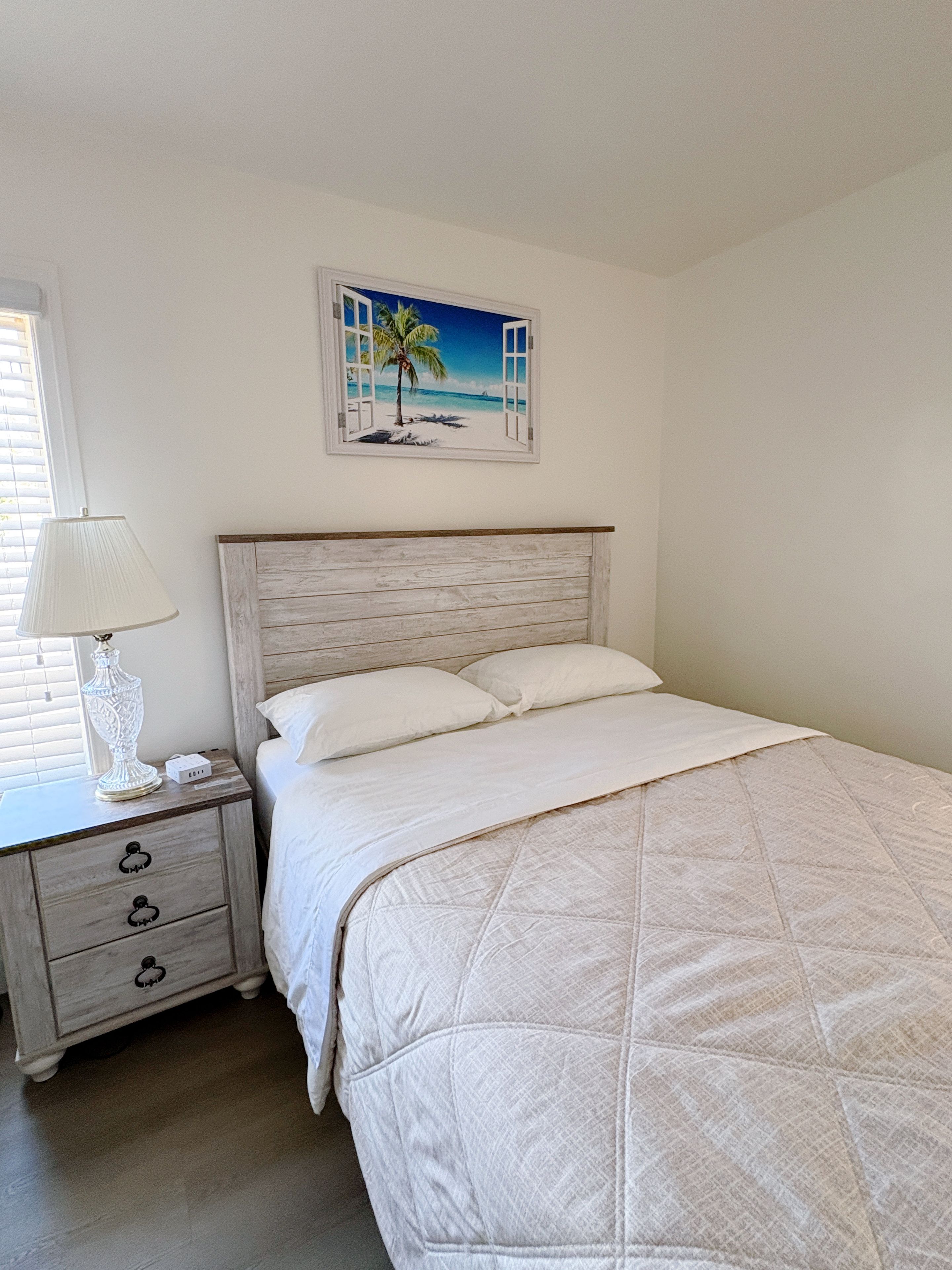 A bedroom with a bed, a nightstand, a lamp, and a painting of a beach scene.