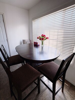 A dining area with a round table, four chairs, a vase with flowers, and a window with blinds. - 2BR Home Near Disneyland & LA - Free Parking (pico Rivera)