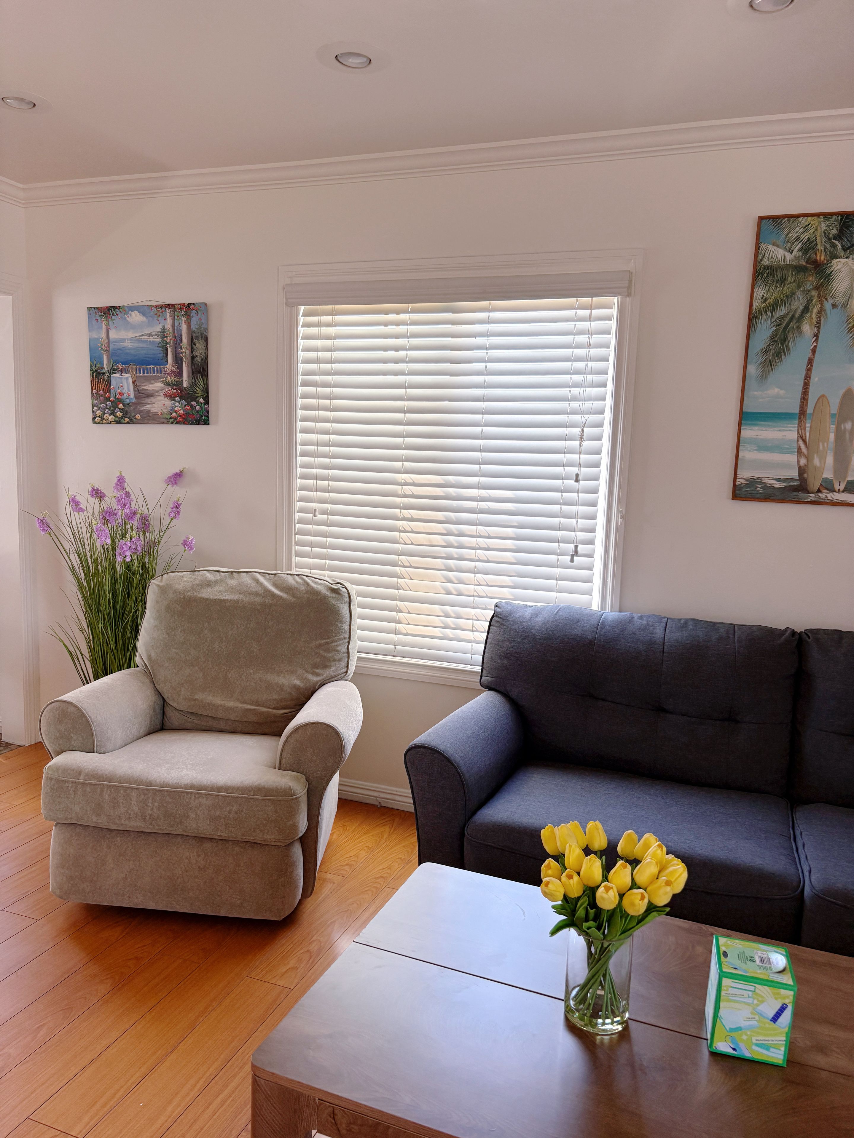 A living room with a beige armchair, a dark grey sofa, a wooden coffee table with a vase of yellow flowers, and a painting of a beach scene.
