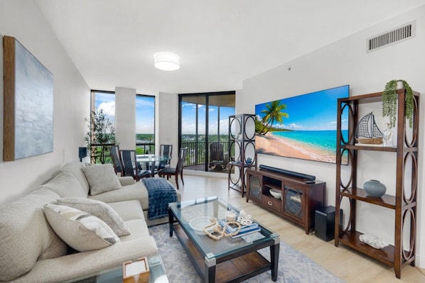 Ein modernes Wohnzimmer mit einer Couch, einem Couchtisch und einem Fernseher. Ein Balkon bietet Blick auf den Strand.
