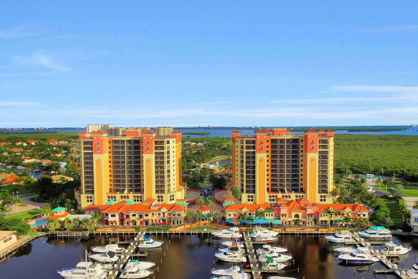 A marina with multiple boats docked, surrounded by two tall buildings with balconies, and a clear view of the water and greenery in the background.