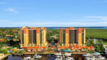 A marina with multiple boats docked, surrounded by two tall buildings with balconies, and a clear view of the water and greenery in the background.
