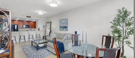 A modern living room with a glass dining table, a sofa, and a kitchen in the background.