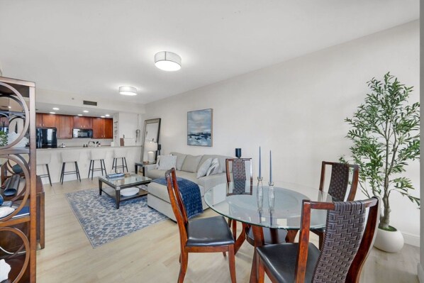 A modern living room with a glass dining table, a sofa, and a kitchen in the background.